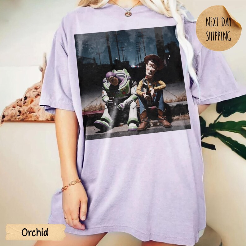 Pode incluir: Camiseta cor de orqu&iacute;dea com um gr&aacute;fico de Buzz Lightyear e Woody de Toy Story. A imagem mostra os personagens sentados juntos em uma cena sombria. A camisa tem um corte folgado. O texto "Next Day Shipping" est&aacute; na camisa.