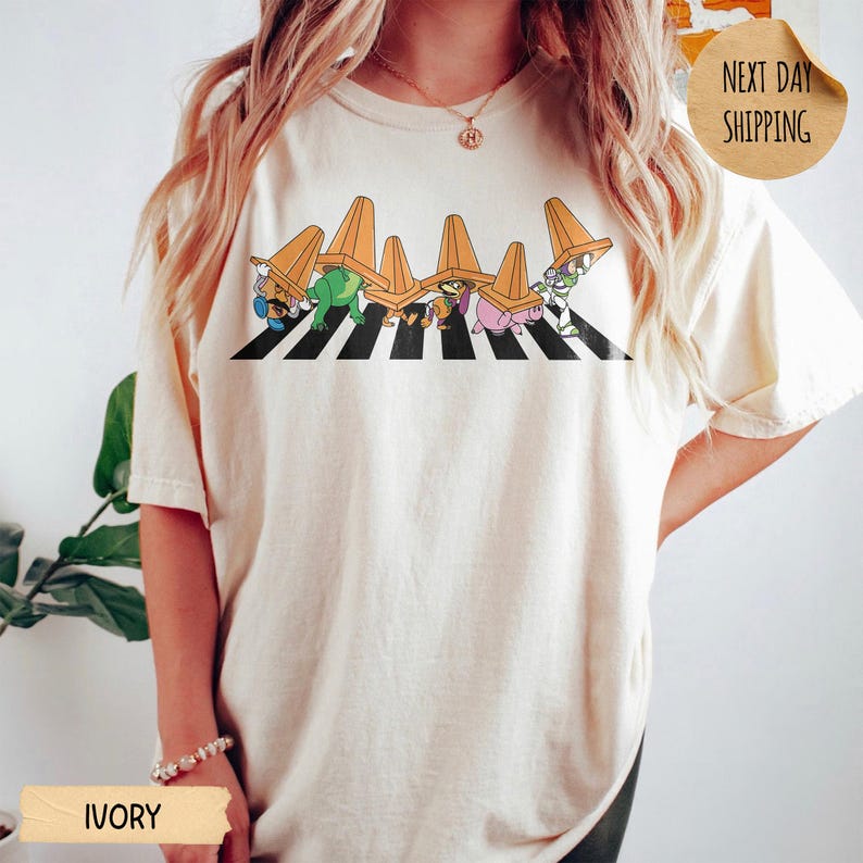May include: An ivory short-sleeve t-shirt featuring a graphic of six cartoon characters walking across a black and white striped zebra crossing, each wearing an orange traffic cone on their head. The shirt has a crew neckline and a relaxed fit.