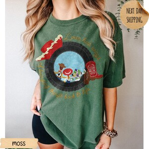 May include: A moss green short-sleeve t-shirt featuring a graphic of a black tire surrounding cartoon characters. The design includes a red hat, a red cowboy boot with the name Taylor, and the text Love has ways of bringing things back to life.