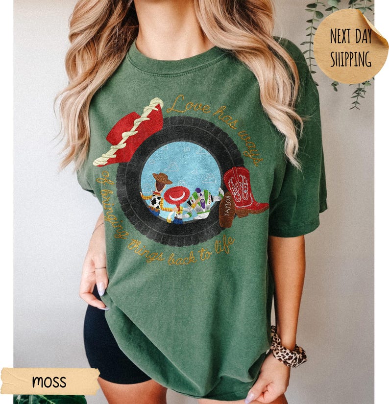 May include: A moss green short-sleeve t-shirt featuring a graphic of a black tire surrounding cartoon characters. The design includes a red hat, a red cowboy boot with the name Taylor, and the text Love has ways of bringing things back to life.