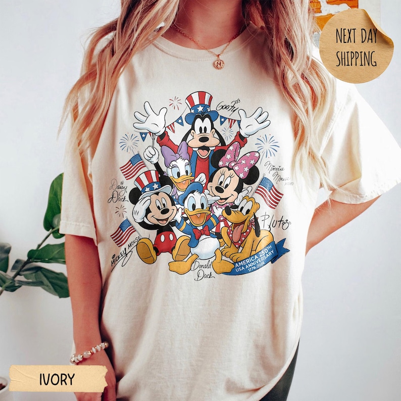 May include: An ivory t-shirt featuring a graphic print of Disney characters including Mickey Mouse, Minnie Mouse, Donald Duck, Daisy Duck, Goofy, and Pluto. The design includes American flags, fireworks, and the text America 250th USA Anniversary.