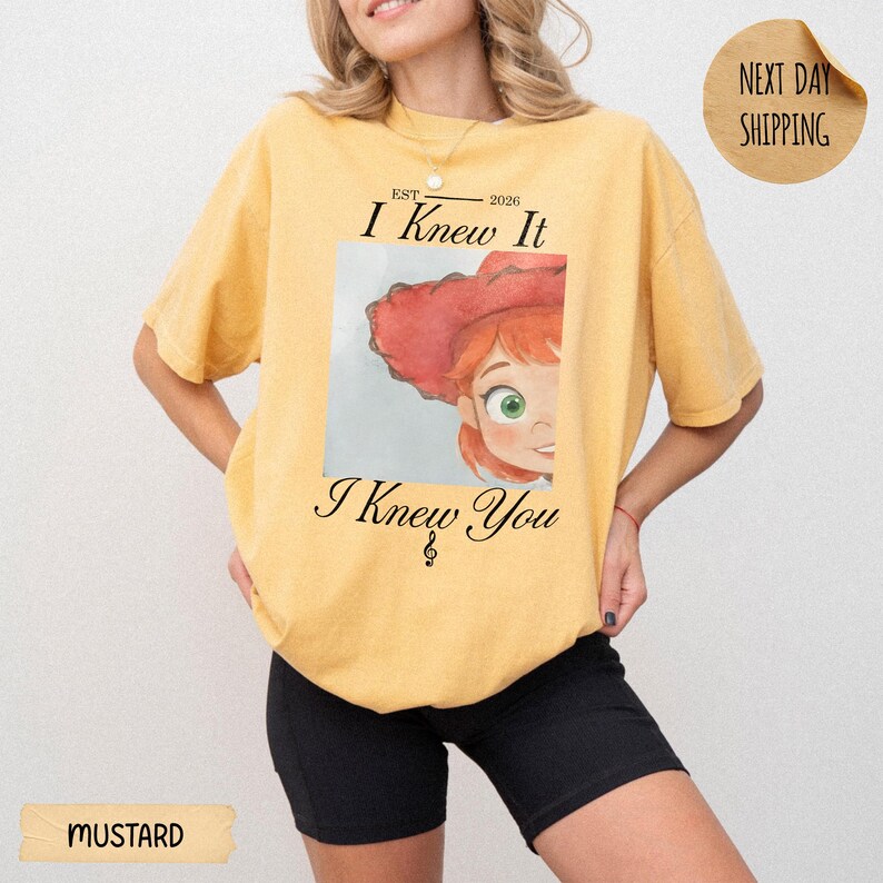 May include: A mustard yellow short-sleeve t-shirt featuring a graphic of a character with red hair and a red hat. The text reads "EST 2026 I Knew It I Knew You" with a small musical note symbol below the text. The shirt has a crew neckline.