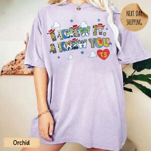 May include: A light purple short-sleeve t-shirt featuring a graphic print with the text I KNEW IT, I KNEW YOU and a red heart with the letters TS. The letters are filled with patterns inspired by cowboy and space ranger character costumes.