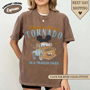 K&ouml;nnte beinhalten: Braunes T-Shirt mit dem Text "Happier than a TORNADO in a trailer park" und einem Cartoon-Abschleppwagen-Design. Das Shirt hat auch den Text "Comfort Colors Espresso" und "Next Day Shipping".