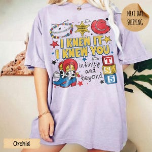 May include: A light purple t-shirt featuring a graphic print with the text I KNEW IT I KNEW YOU infinity and beyond. The design includes a colorful beaded bracelet, a yellow sheriff star, a red cowboy hat, cowboy boots, and three alphabet blocks.