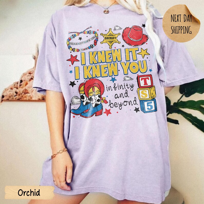 May include: A light purple t-shirt featuring a graphic print with the text I KNEW IT I KNEW YOU infinity and beyond. The design includes a colorful beaded bracelet, a yellow sheriff star, a red cowboy hat, cowboy boots, and three alphabet blocks.