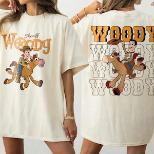 May include: Cream-colored t-shirt featuring a cartoon image of Sheriff Woody riding a horse. The front of the shirt has the text "Sheriff Woody" in orange and brown. The back of the shirt has the word "Woody" repeated multiple times.