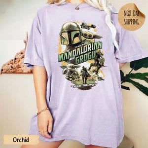 Pu&ograve; includere: T-shirt color orchidea con una grafica della serie Star Wars, "The Mandalorian and Grogu". Il design include immagini di The Mandalorian, Grogu e il testo "This is the Way."