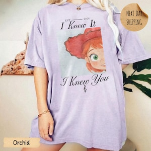 May include: A light purple short-sleeve t-shirt featuring a graphic of a cartoon character with red hair and a red hat. The text above reads "EST. 2026 I Knew It" and the text below reads "I Knew You" with a small musical note symbol.