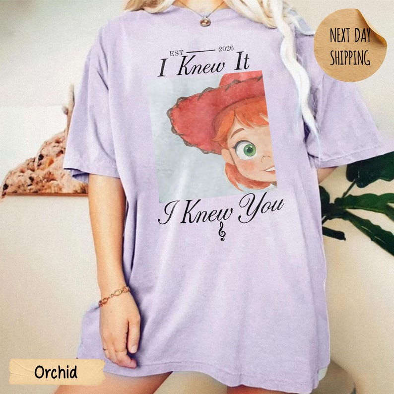 May include: A light purple short-sleeve t-shirt featuring a graphic of a cartoon character with red hair and a red hat. The text above reads "EST. 2026 I Knew It" and the text below reads "I Knew You" with a small musical note symbol.