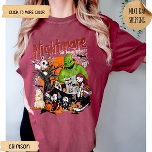 May include: A crimson short-sleeve t-shirt featuring a graphic print with the text Nightmare on Main Street. The design includes characters like Jack Skellington, Sally, and Oogie Boogie, along with a tombstone, a pumpkin, and a spider web.