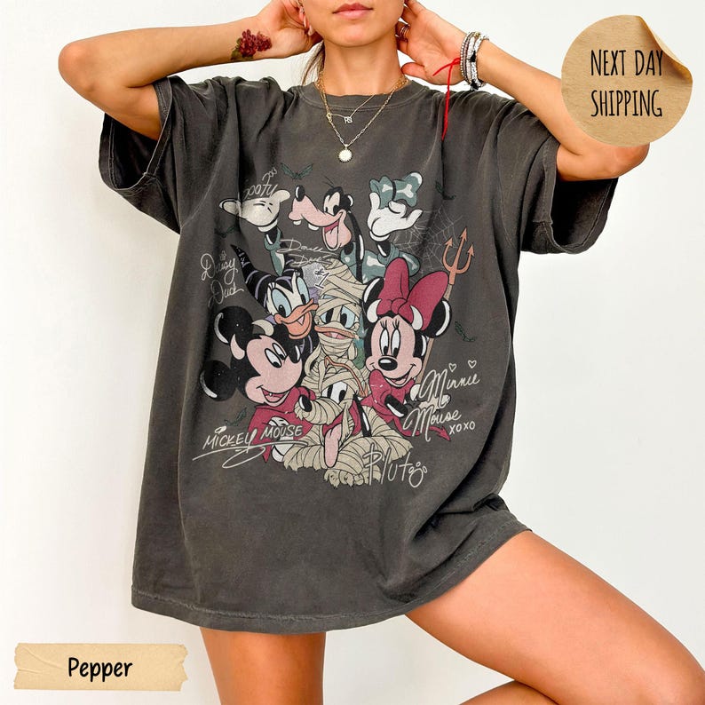 Peut inclure: T-shirt gris fonc&eacute; avec un motif d'Halloween de personnages Disney, dont Mickey et Minnie Mouse, Donald Duck et Dingo. Le t-shirt porte les inscriptions "Mickey Mouse" et "Minnie Mouse" en cursive.