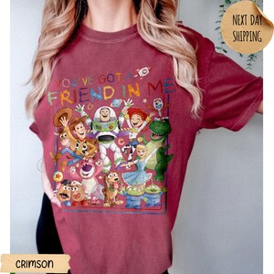 May include: A crimson short-sleeve t-shirt featuring a colorful graphic print of various Toy Story characters including Woody, Buzz Lightyear, and Jessie. The text "YOU'VE GOT A FRIEND IN ME" is printed in multicolored letters above the character group.