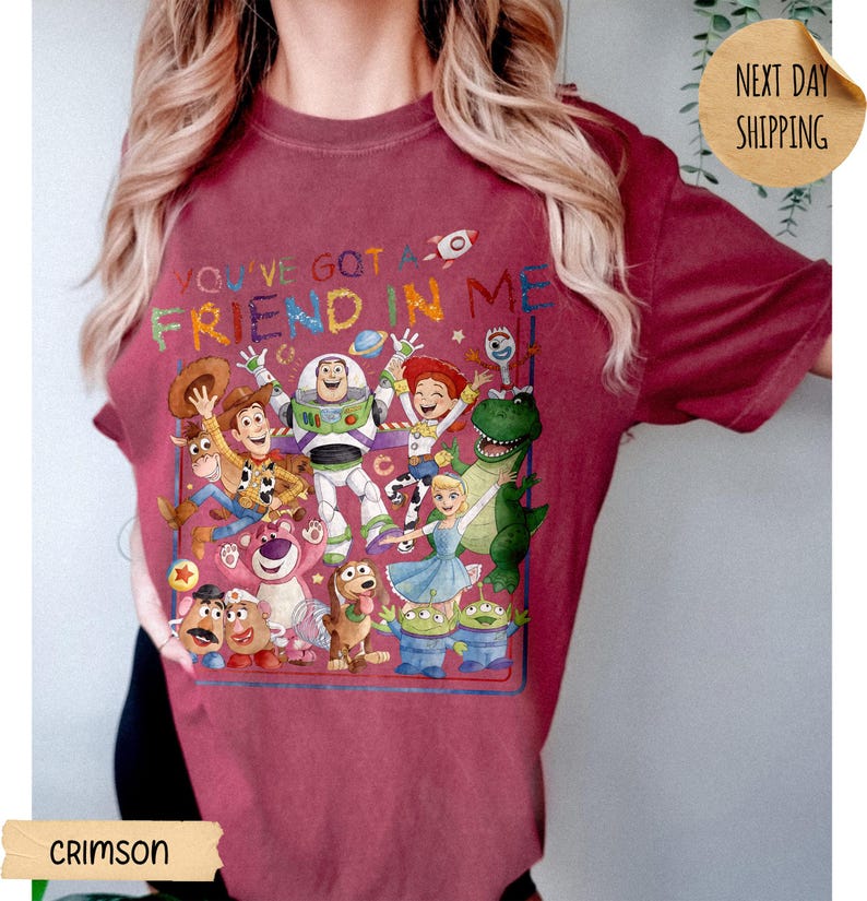May include: A crimson short-sleeve t-shirt featuring a colorful graphic print of various Toy Story characters including Woody, Buzz Lightyear, and Jessie. The text "YOU'VE GOT A FRIEND IN ME" is printed in multicolored letters above the character group.