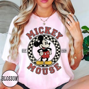 Puede incluir: Camiseta rosa claro con un dise&ntilde;o vintage de Mickey Mouse. El dise&ntilde;o incluye las palabras "Mickey Mouse" en una fuente retro, con "Est. 1928" y un c&iacute;rculo a cuadros alrededor de Mickey. La palabra "Blossom" tambi&eacute;n est&aacute; impresa en la camiseta.