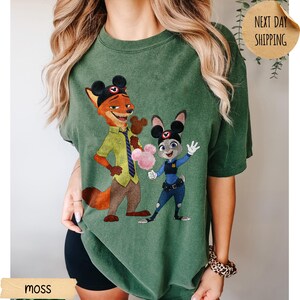 May include: Moss green t-shirt featuring a cartoon illustration of Nick Wilde and Judy Hopps from Zootopia. The characters are wearing Mickey Mouse ears and holding cotton candy. The shirt has a relaxed fit.