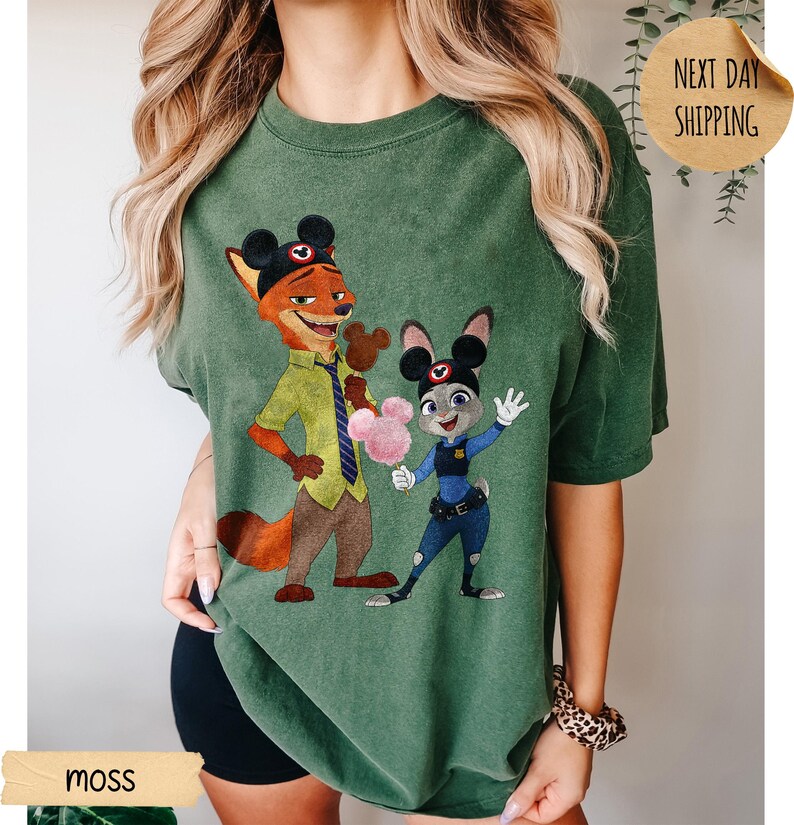 May include: Moss green t-shirt featuring a cartoon illustration of Nick Wilde and Judy Hopps from Zootopia. The characters are wearing Mickey Mouse ears and holding cotton candy. The shirt has a relaxed fit.