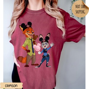May include: Crimson-colored t-shirt featuring a cartoon illustration of Nick Wilde and Judy Hopps from Zootopia. Nick is holding a Mickey Mouse-shaped ice cream, and Judy is holding cotton candy. The shirt also has the text "CRIMSON" and "NEXT DAY SHIPPING".