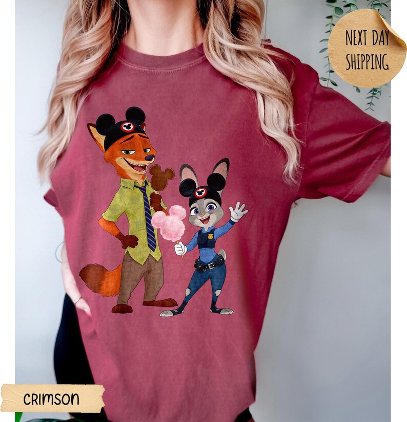 May include: Crimson-colored t-shirt featuring a cartoon illustration of Nick Wilde and Judy Hopps from Zootopia. Nick is holding a Mickey Mouse-shaped ice cream, and Judy is holding cotton candy. The shirt also has the text "CRIMSON" and "NEXT DAY SHIPPING".