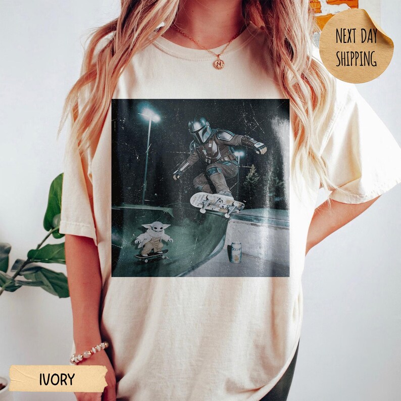 May include: Ivory-colored t-shirt featuring a graphic of The Mandalorian skateboarding, with Grogu also on a skateboard. The image is set in a skate park. The shirt also has the text "Next Day Shipping".