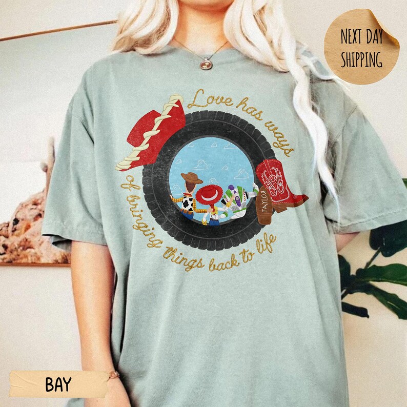 May include: A sage green t-shirt featuring a graphic of a black tire framing a scene with cartoon characters. The design includes a red cowboy hat, a red cowboy boot, and the text "Love has ways of bringing things back to life" in gold script.