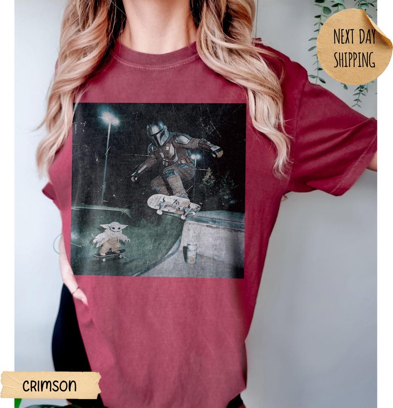 May include: Crimson-colored t-shirt featuring a graphic of The Mandalorian skateboarding, with Grogu also on a skateboard. The image has a distressed, vintage look. The shirt is a solid color with a crew neck.