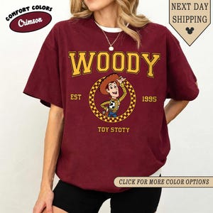 Puede incluir: Camiseta de color carmes&iacute; con la palabra "WOODY" en amarillo, sobre una imagen de dibujos animados de Woody de Toy Story. La camiseta tambi&eacute;n incluye el texto "EST 1995" y "TOY STOTY". Parte de la colecci&oacute;n Comfort Colors.