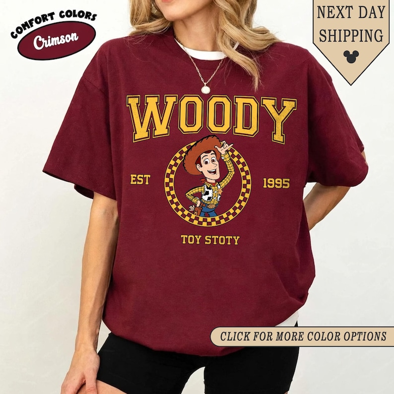 Puede incluir: Camiseta de color carmes&iacute; con la palabra "WOODY" en amarillo, sobre una imagen de dibujos animados de Woody de Toy Story. La camiseta tambi&eacute;n incluye el texto "EST 1995" y "TOY STOTY". Parte de la colecci&oacute;n Comfort Colors.