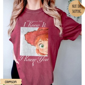 May include: A crimson short-sleeve t-shirt featuring a graphic print of a cartoon character with red hair and a red hat. The text reads EST 2026, I Knew It, and I Knew You with a treble clef symbol below the text. The design is centered on the chest.