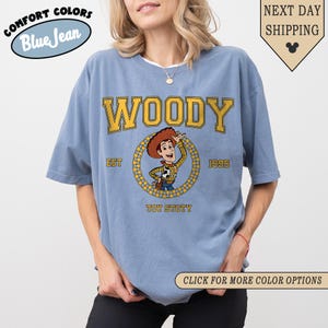 Puede incluir: Una camiseta azul claro con la palabra "WOODY" en letras amarillas. La camiseta presenta una imagen de dibujos animados de Woody de Toy Story, con el texto "EST 1995" y "TOY STOTY". La camiseta tambi&eacute;n tiene el texto "COMFORT COLORS Blue Jean".