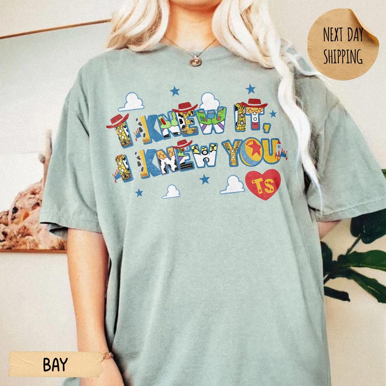 May include: A sage green short-sleeve t-shirt featuring the text I KNEW IT, I KNEW YOU in a colorful, patterned font inspired by animated characters. The design includes small blue stars, white clouds, and a red heart with the letters TS inside.