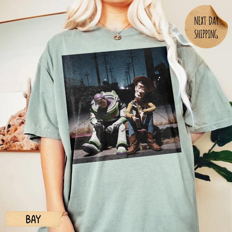 Pode incluir: T-shirt verde claro com uma imagem desgastada de Buzz Lightyear e Woody de Toy Story sentados numa cal&ccedil;ada. A imagem tem uma qualidade escura e cinematogr&aacute;fica, com postes de luz ao fundo. A t-shirt tem um corte folgado.