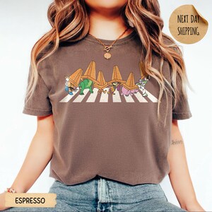 May include: A dark brown short-sleeve t-shirt featuring a graphic of five cartoon characters walking across a white striped zebra crossing, each wearing an orange traffic cone on their head. The shirt has a crew neckline and a relaxed fit.