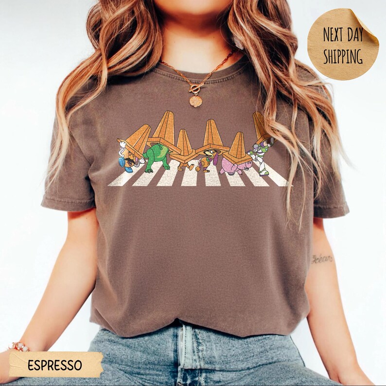 May include: A dark brown short-sleeve t-shirt featuring a graphic of five cartoon characters walking across a white striped zebra crossing, each wearing an orange traffic cone on their head. The shirt has a crew neckline and a relaxed fit.