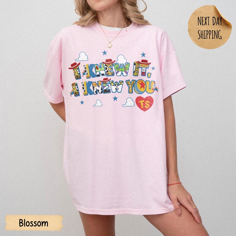 May include: A light pink short-sleeve t-shirt featuring the text I KNEW IT, I KNEW YOU in a multi-colored block font with character patterns. The design includes small blue stars, white cloud outlines, and a red heart with the letters TS inside.