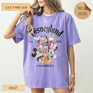 Op de afbeelding: Violetkleurig T-shirt met een Disneyland-afbeelding. Het ontwerp bevat het Disneyland-logo, de tekst "Est 1955 California" en stripfiguren zoals Mickey Mouse, Minnie Mouse, Donald Duck en Goofy.
