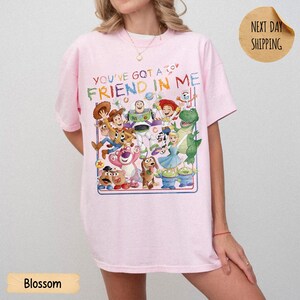 May include: A light pink short-sleeve t-shirt featuring a colorful graphic print of various animated characters from the Toy Story franchise. The text above the characters reads, YOU'VE GOT A FRIEND IN ME, in a multi-colored, playful font style.