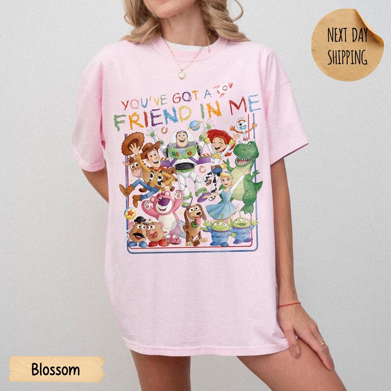 May include: A light pink short-sleeve t-shirt featuring a colorful graphic print of various animated characters from the Toy Story franchise. The text above the characters reads, YOU'VE GOT A FRIEND IN ME, in a multi-colored, playful font style.