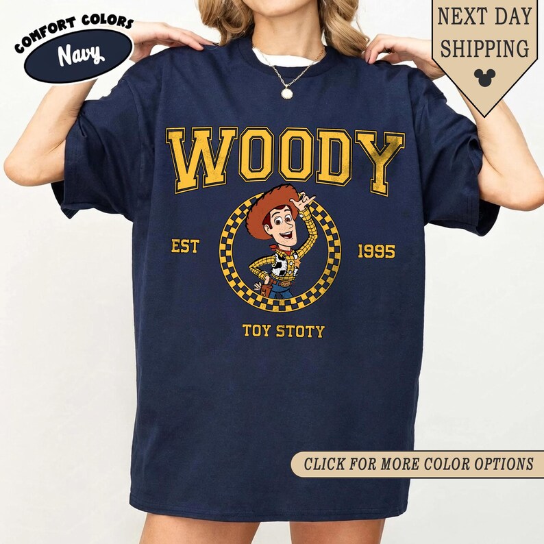 Puede incluir: Camiseta azul marino con la palabra "WOODY" en letras doradas. Debajo, un gr&aacute;fico circular de un vaquero de dibujos animados, con "EST 1995" y "TOY STOTY". La camiseta tiene un corte holgado y es c&oacute;moda.