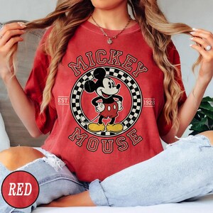 Puede incluir: Camiseta roja con un dise&ntilde;o vintage de Mickey Mouse. El dise&ntilde;o incluye un marco circular con un patr&oacute;n de cuadros, las palabras "Mickey Mouse" y el a&ntilde;o "1928". La camiseta es de color rojo liso.