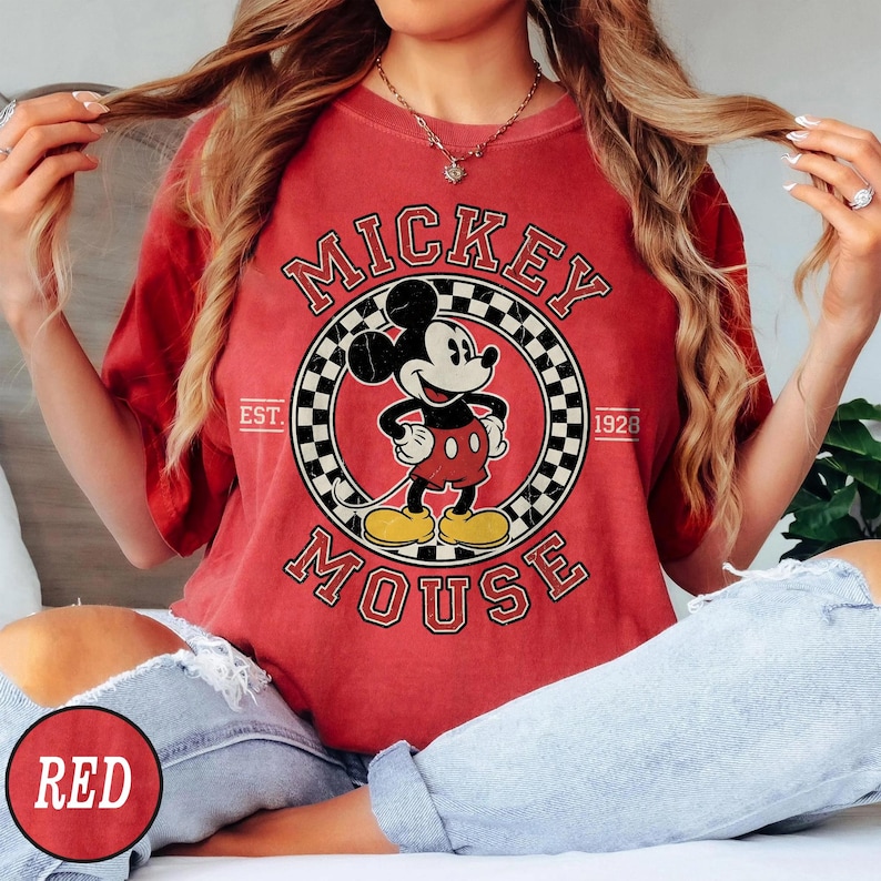 Puede incluir: Camiseta roja con un dise&ntilde;o vintage de Mickey Mouse. El dise&ntilde;o incluye un marco circular con un patr&oacute;n de cuadros, las palabras "Mickey Mouse" y el a&ntilde;o "1928". La camiseta es de color rojo liso.