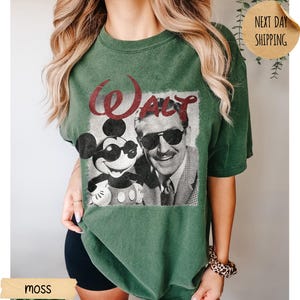 May include: Moss green t-shirt featuring a graphic of Mickey Mouse wearing sunglasses and Walt Disney. The word "Walt" is printed in red above the image. The shirt is a relaxed fit.