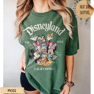 May include: A moss green short-sleeve t-shirt featuring a graphic print of Mickey Mouse, Minnie Mouse, Donald Duck, Daisy Duck, Goofy, and Pluto. The text reads Disneyland, Est 1955, and California, with character names written in script.