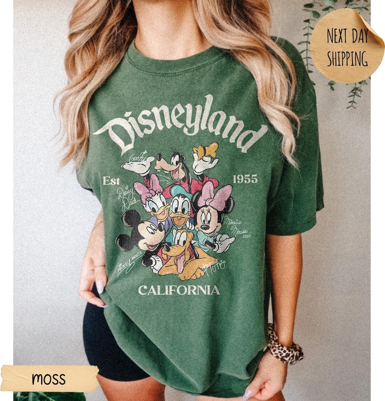 May include: A moss green short-sleeve t-shirt featuring a graphic print of Mickey Mouse, Minnie Mouse, Donald Duck, Daisy Duck, Goofy, and Pluto. The text reads Disneyland, Est 1955, and California, with character names written in script.
