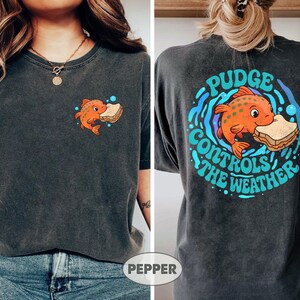 May include: Charcoal grey t-shirt with a cartoon orange fish and sandwich graphic. The front has a small fish design, and the back features a larger design with the text "Pudge Controls the Weather" in a circular arrangement.