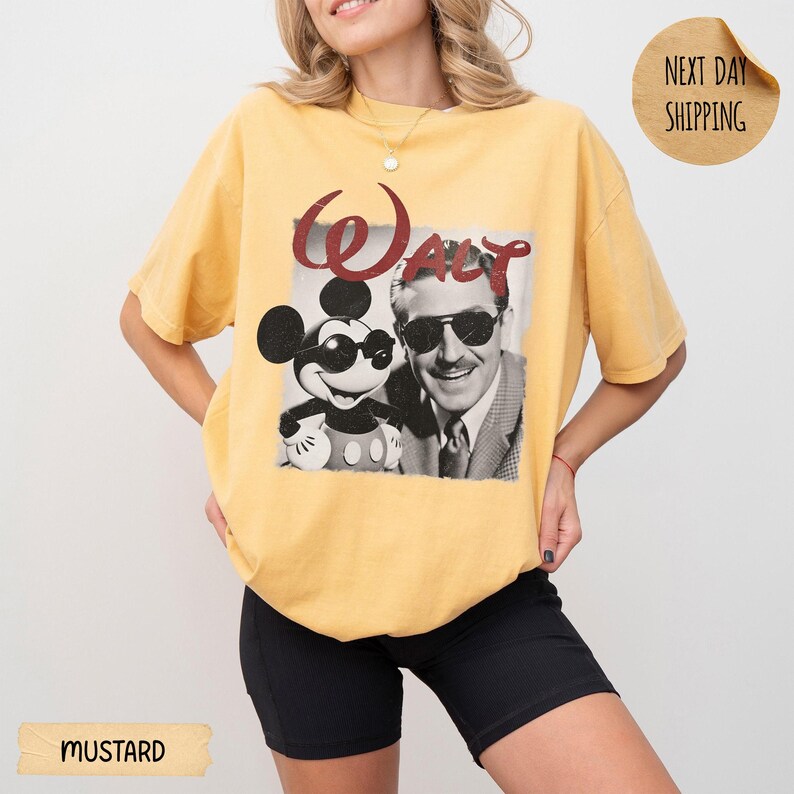 May include: Mustard-colored t-shirt featuring a black and white graphic of Mickey Mouse and Walt Disney with the word "Walt" in red. The shirt has short sleeves and a relaxed fit.