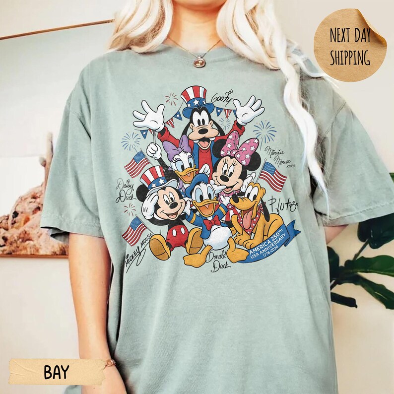 May include: A sage green t-shirt featuring a graphic print of Mickey Mouse, Donald Duck, Goofy, Daisy Duck, Minnie Mouse, and Pluto. The characters are wearing patriotic attire with American flags and fireworks. Text includes character names and America 250th USA Anniversary 1776-2026.