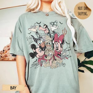 Peut inclure: T-shirt vert clair avec un motif d'Halloween de personnages Disney, dont Mickey, Minnie, Donald et Dingo. Le design comprend des chauves-souris, des toiles d'araign&eacute;es et des signatures de personnages. Le t-shirt a une coupe ample.