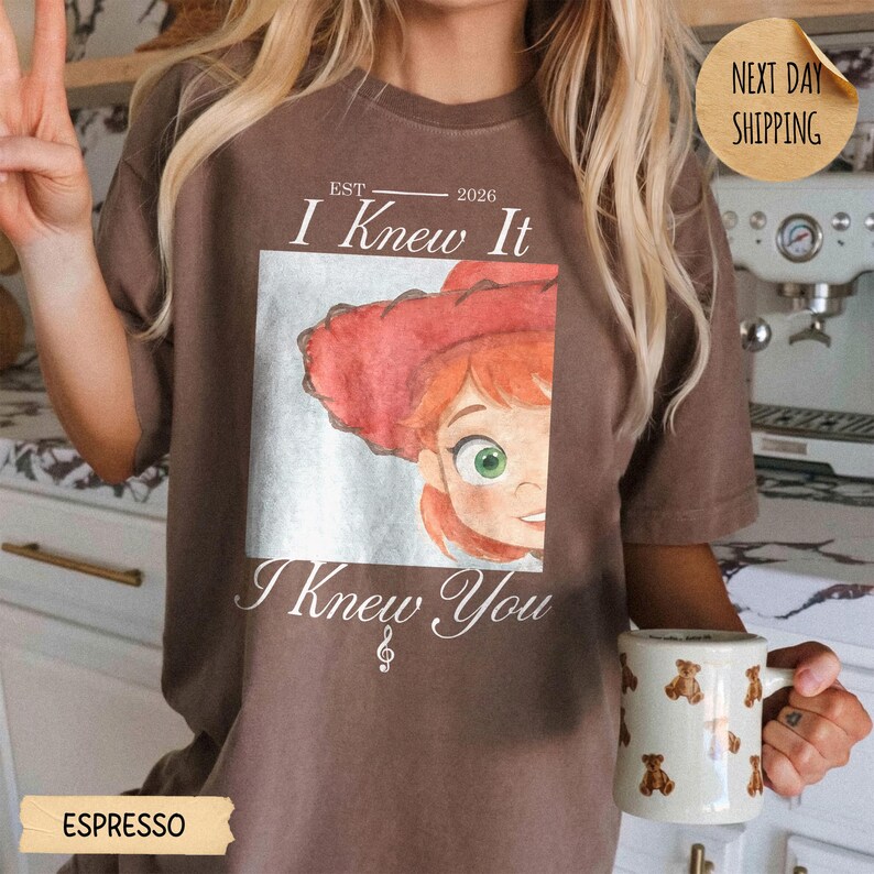 May include: A brown t-shirt featuring a square graphic of a cartoon character with red hair and a red hat. The text reads EST 2026 I Knew It I Knew You with a treble clef symbol. The shirt is worn with a white mug patterned with small brown teddy bears.