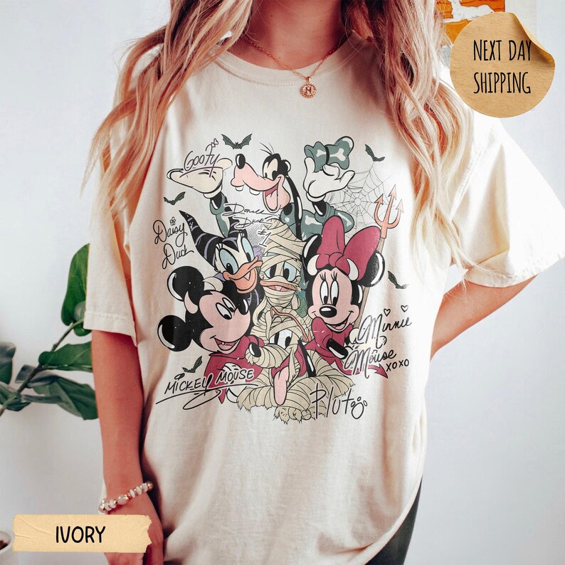 Peut inclure: T-shirt ivoire avec un motif d'Halloween de personnages Disney, dont Mickey, Minnie, Donald et Dingo. Le motif comprend des chauves-souris, des toiles d'araign&eacute;es et des signatures de personnages. Le t-shirt a une coupe d&eacute;contract&eacute;e.